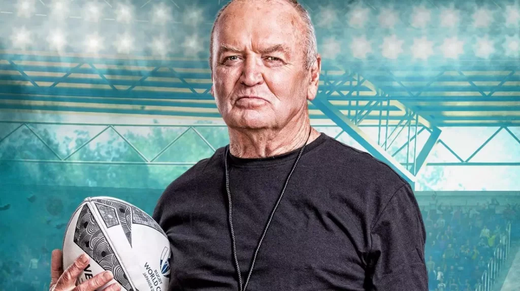 Sir Graham Henry, legendary former All Blacks coach, holding a rugby ball in a stadium setting.