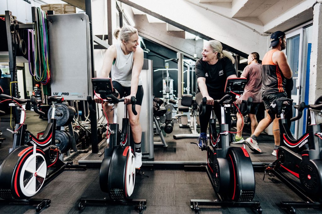 Two women smiling and training together on spin bikes at Body Torque Gym in Newmarket, Auckland.
