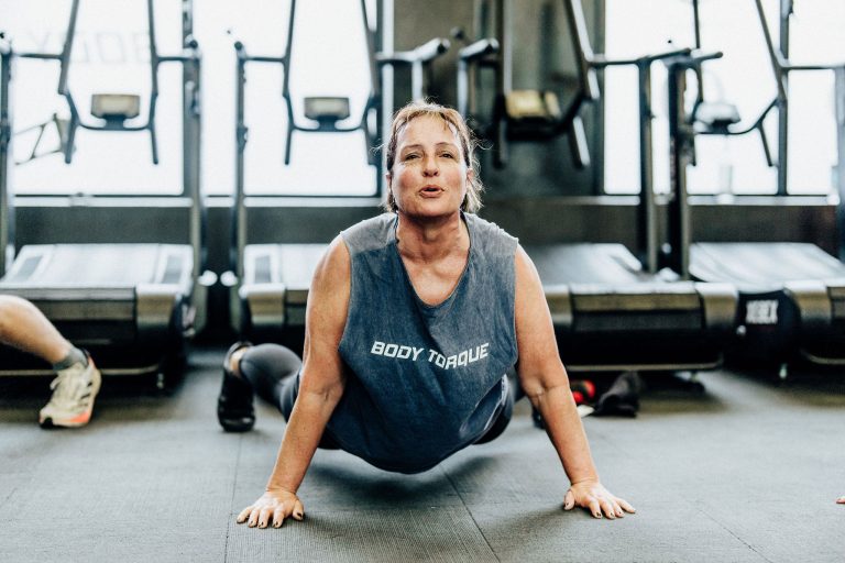 Middle-aged woman in a Body Torque Gym T-shirt performing a floor stretch exercise at Body Torque Gym in Newmarket, Auckland.
