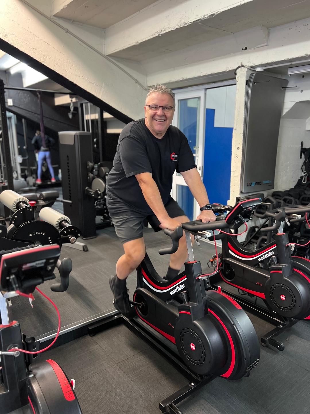 Middle-aged man smiling while cycling on a Watt bike in Body Torque gym as part of his fitness transformation journey.